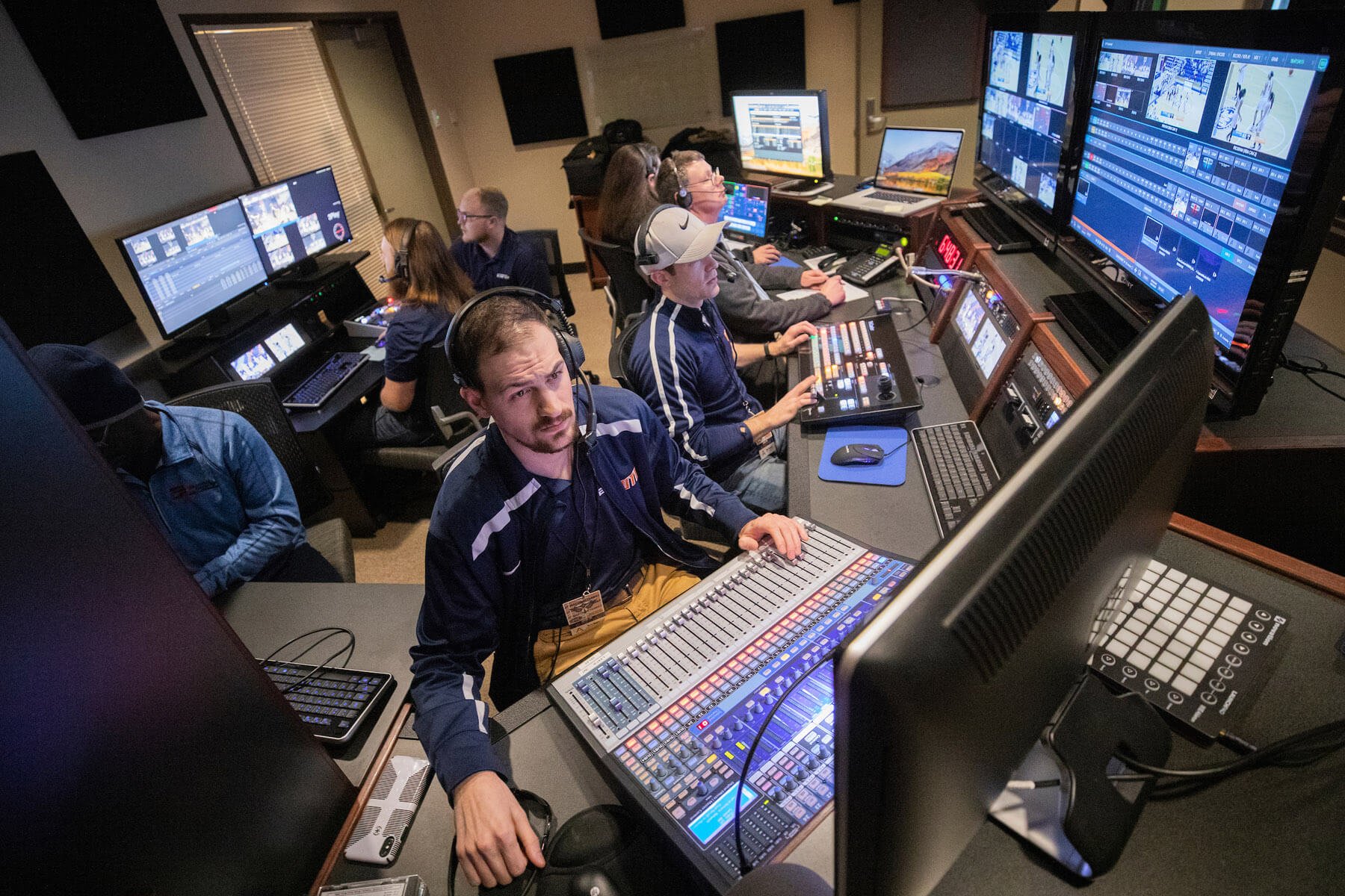 Broadcast control room with technicians working at multiple screens and sound mixing consoles, wearing headphones and navy uniforms.